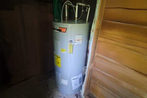 %Focuskw% | Get Lit Electrical, Plumbing &Amp; Hvac A gray water heater is installed in a small utility space with wooden walls and exposed pipes connected at the top.