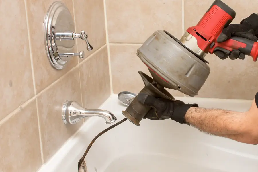 A person uses a power drain snake to clear a clogged bathtub drain in a tiled bathroom.