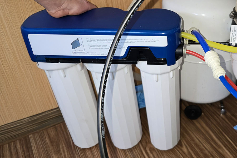 A person’s hand is adjusting a three-stage water filtration system installed under a sink, with tubes connected to the unit.