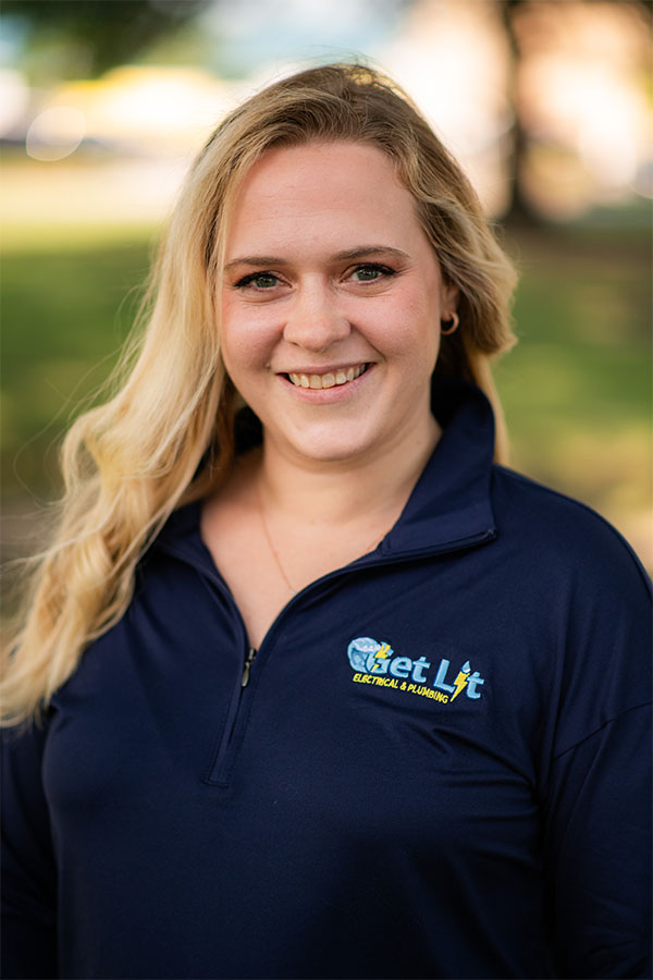 A woman with long blonde hair smiles at the camera, wearing a navy blue "Get Lit Electrical & Lighting" shirt, standing outdoors.