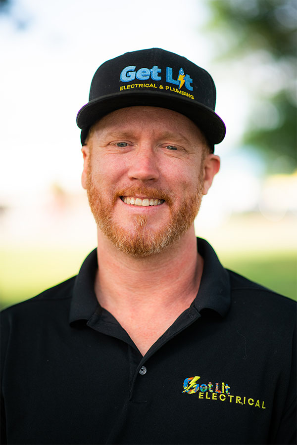 A man with a red beard wearing a black "Get Lit Electrical & Plumbing" cap and polo shirt stands outdoors, smiling at the camera.
