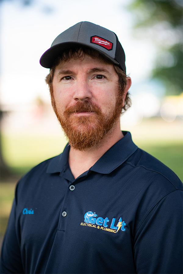 Man with a reddish beard wears a black Milwaukee hat and a navy Get Lit Electrical & Plumbing polo, standing outdoors with trees and blurred background.