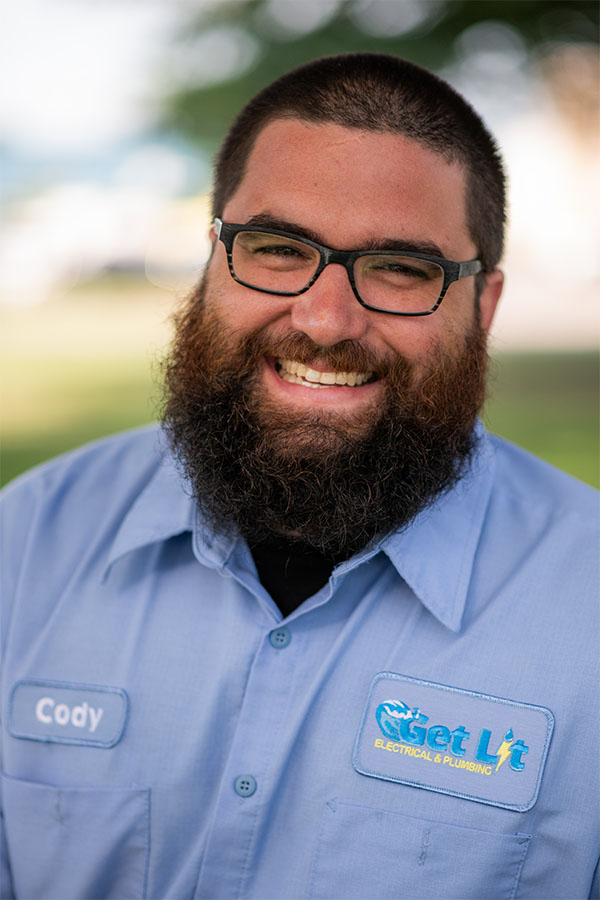 A man with a beard and glasses, wearing a light blue "Get Lit Electrical & Plumbing" shirt with a name tag that says "Cody," smiles at the camera outdoors.