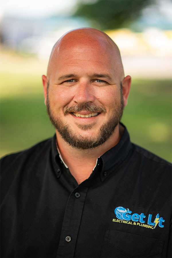 A smiling man with a beard wearing a black "Get Lit Electrical & Plumbing" shirt, standing outdoors with a blurred green and blue background.