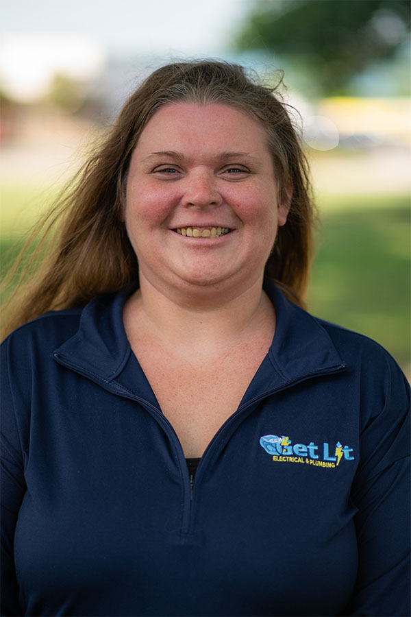 A woman with long hair smiles outdoors, wearing a navy blue zip-up shirt with the "Get Lit Electrical & Plumbing" logo.