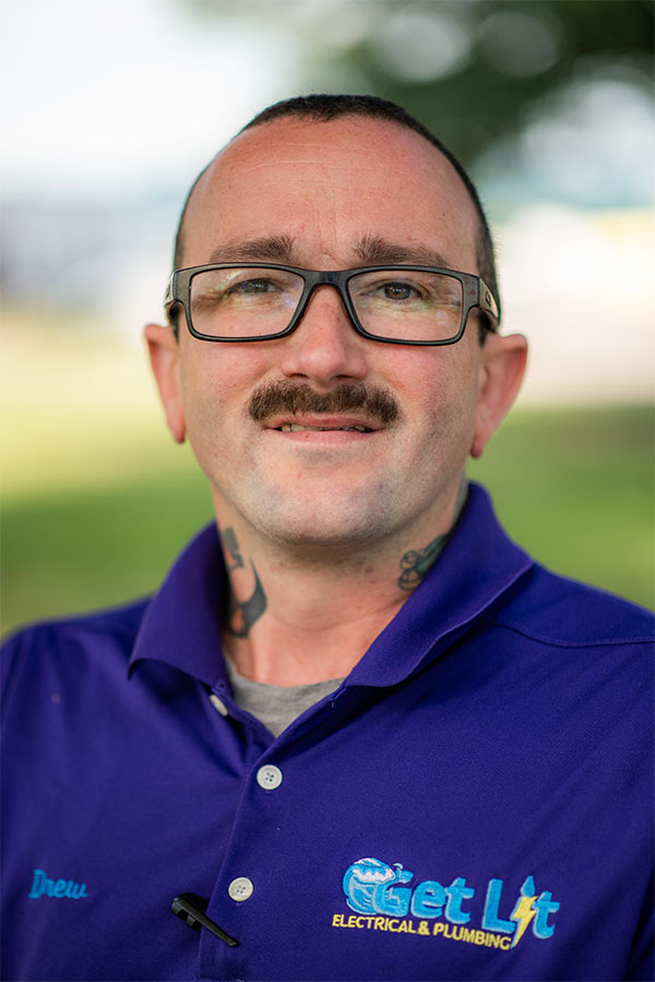 A man with glasses and a mustache wears a purple "Get Lit Electrical & Plumbing" shirt and stands outdoors, looking at the camera.