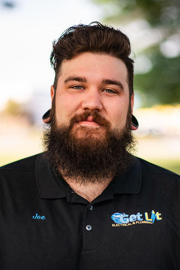 A man with a beard and stretched earlobes wears a black "Get Lit Electrical & Plumbing" polo shirt with the name "Joe" embroidered on it, standing outdoors.