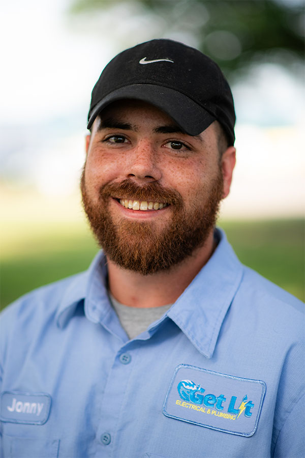 A man with a beard wearing a black cap and a blue "Get Lit Electrical & Plumbing" work shirt stands outdoors, smiling at the camera.
