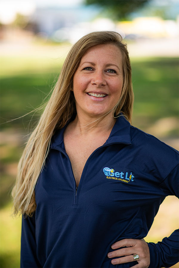 A woman with long blonde hair stands outdoors, smiling, wearing a navy zip-up shirt with the "Get Lit Electrical & Plumbing" logo on the chest.