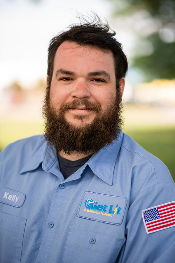 Man with a beard wearing a light blue "Get Lit Electrical & Plumbing" uniform shirt with an American flag patch and a name tag that reads "Kelly," outdoors with greenery in the background.