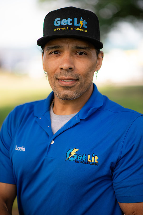 A man wearing a "Get Lit Electrical & Plumbing" hat and shirt with the name "Louis" embroidered, standing outdoors and looking at the camera.