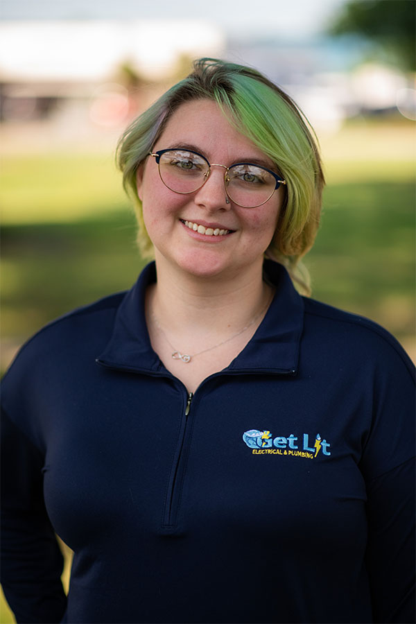 A person with short, light green hair and glasses, wearing a navy "Get Lit Electrical & Plumbing" shirt, stands outdoors and smiles at the camera.