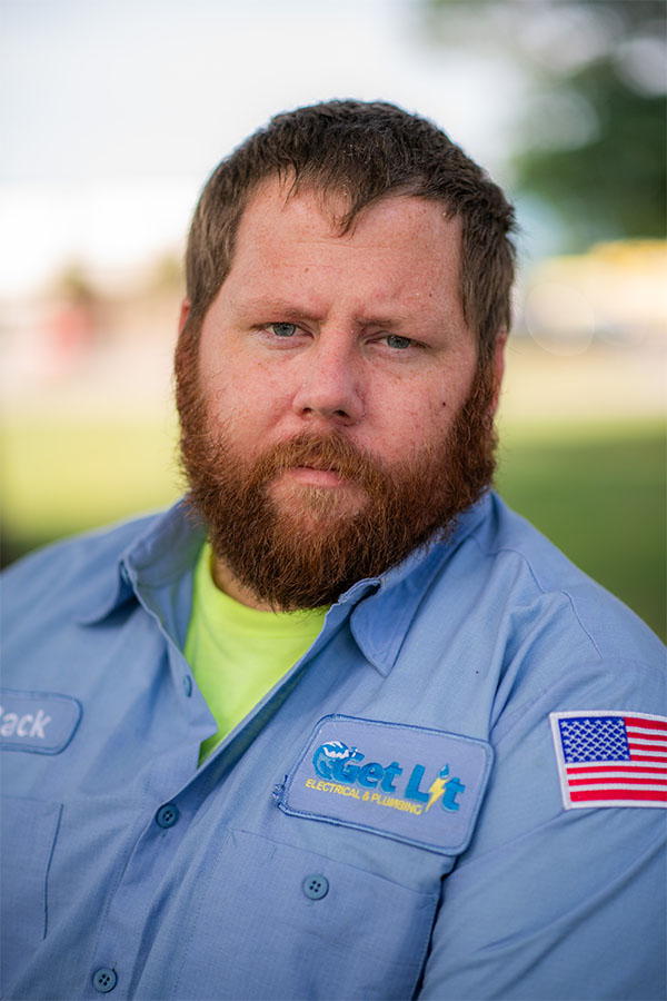 A man with a red beard in a blue work shirt with a US flag patch and "Get Lit Electrical & Plumbing" logo looks at the camera outdoors.