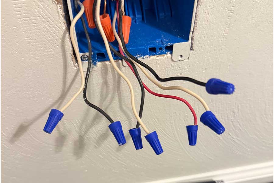 Several electrical wires with blue and orange wire nuts are exposed from an open blue electrical box mounted in a wall.