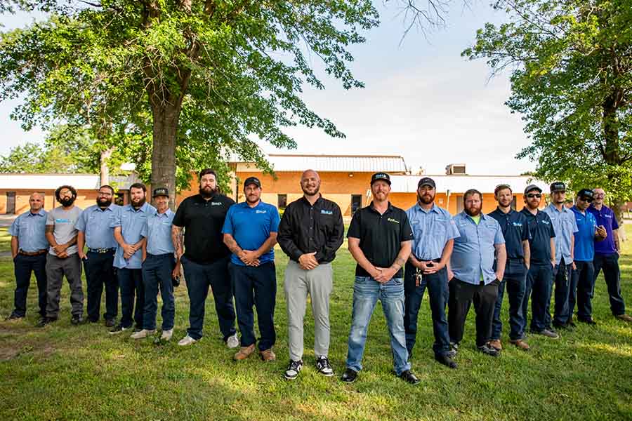 A group of men wearing work uniforms stand outside on grass in front of a building and trees, posing for an about us group photo.