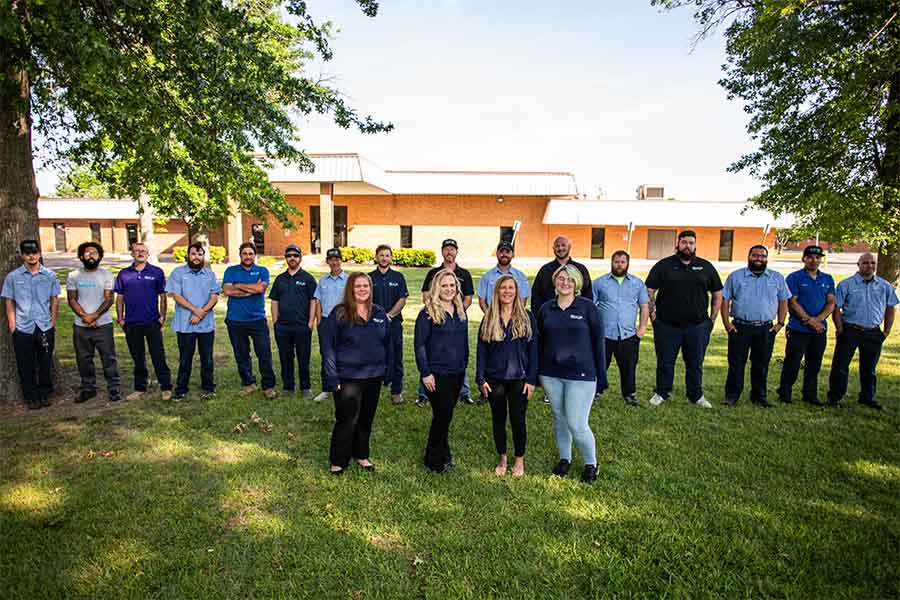A group of eighteen people stands in two rows on grass outside a brick building, with trees and clear sky in the background—capturing the spirit of our team for the about us page.