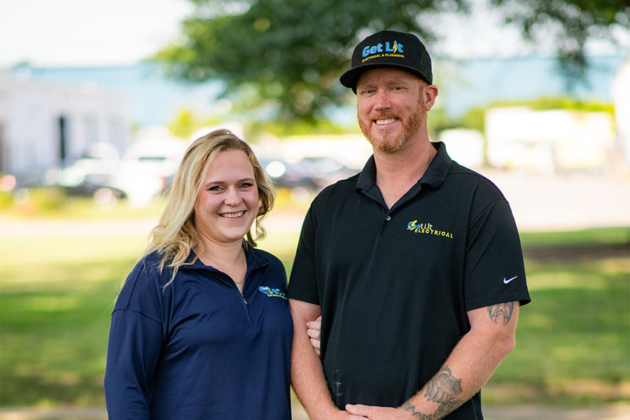Two people stand outdoors smiling in branded shirts and a "Get Lit" hat, embodying the spirit of our team. This candid moment captures the heart of our about us story, with trees and buildings softly blurred in the background.