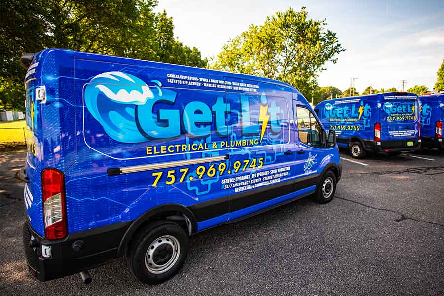 Blue "Get Lit Electrical & Plumbing" service van with contact number parked in a lot, promoting their maintenance plan; another similar van is visible in the background with trees and grass nearby.